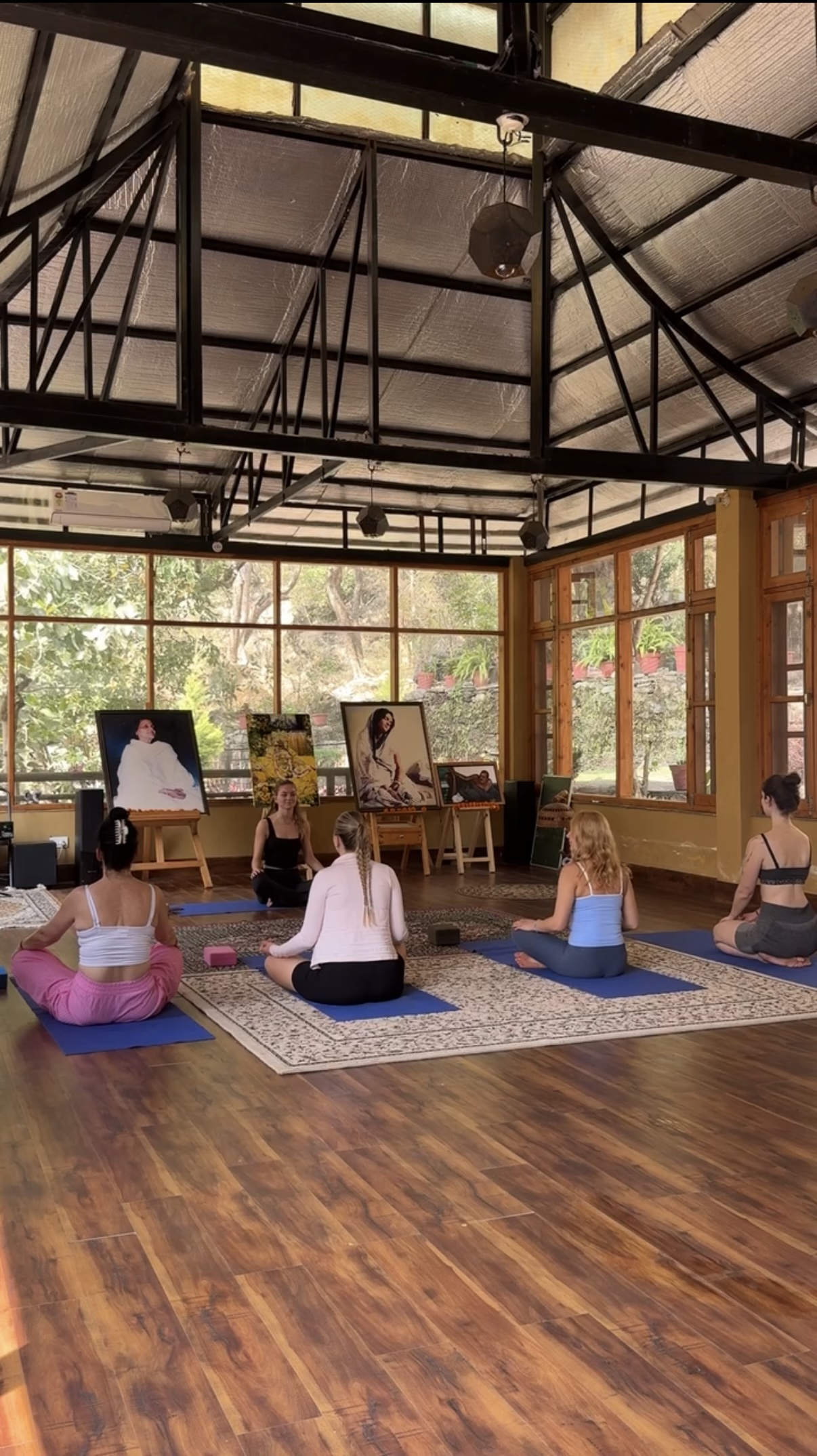 Group of women practicing yoga in a spacious studio with large windows and artwork displayed during yoga retreat in Rishikesh, India. 