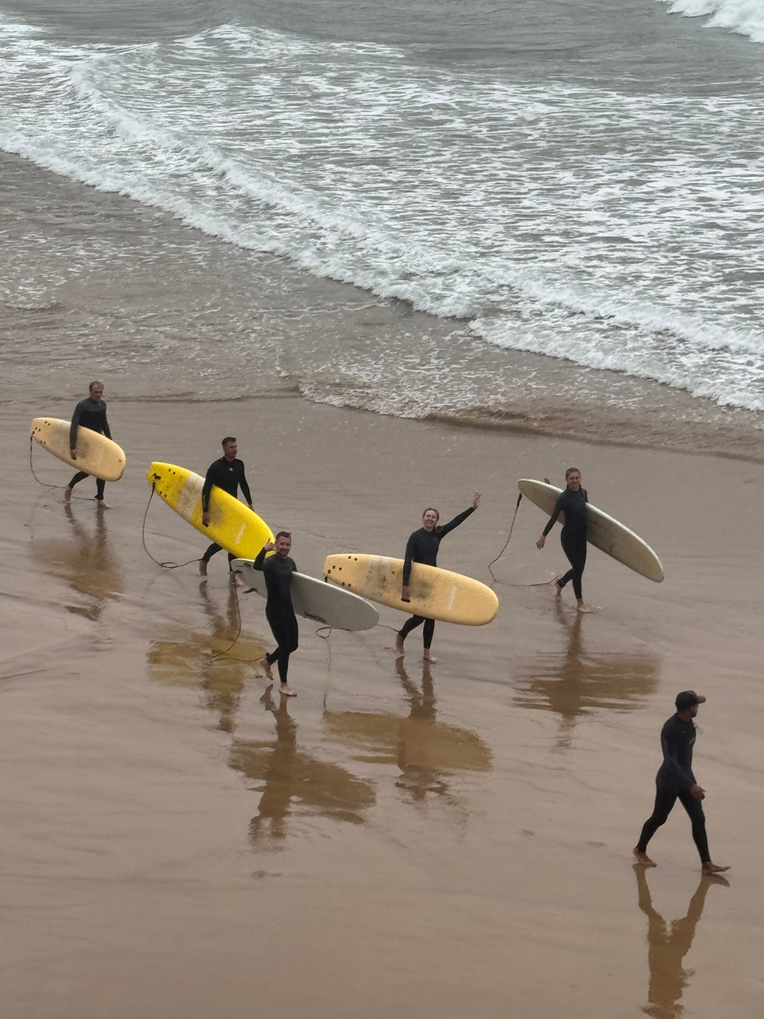 Group of surfers in black wetsuits walking along the shoreline, carrying surfboards, with waves in the background, surfing, Morocco, Imsouane